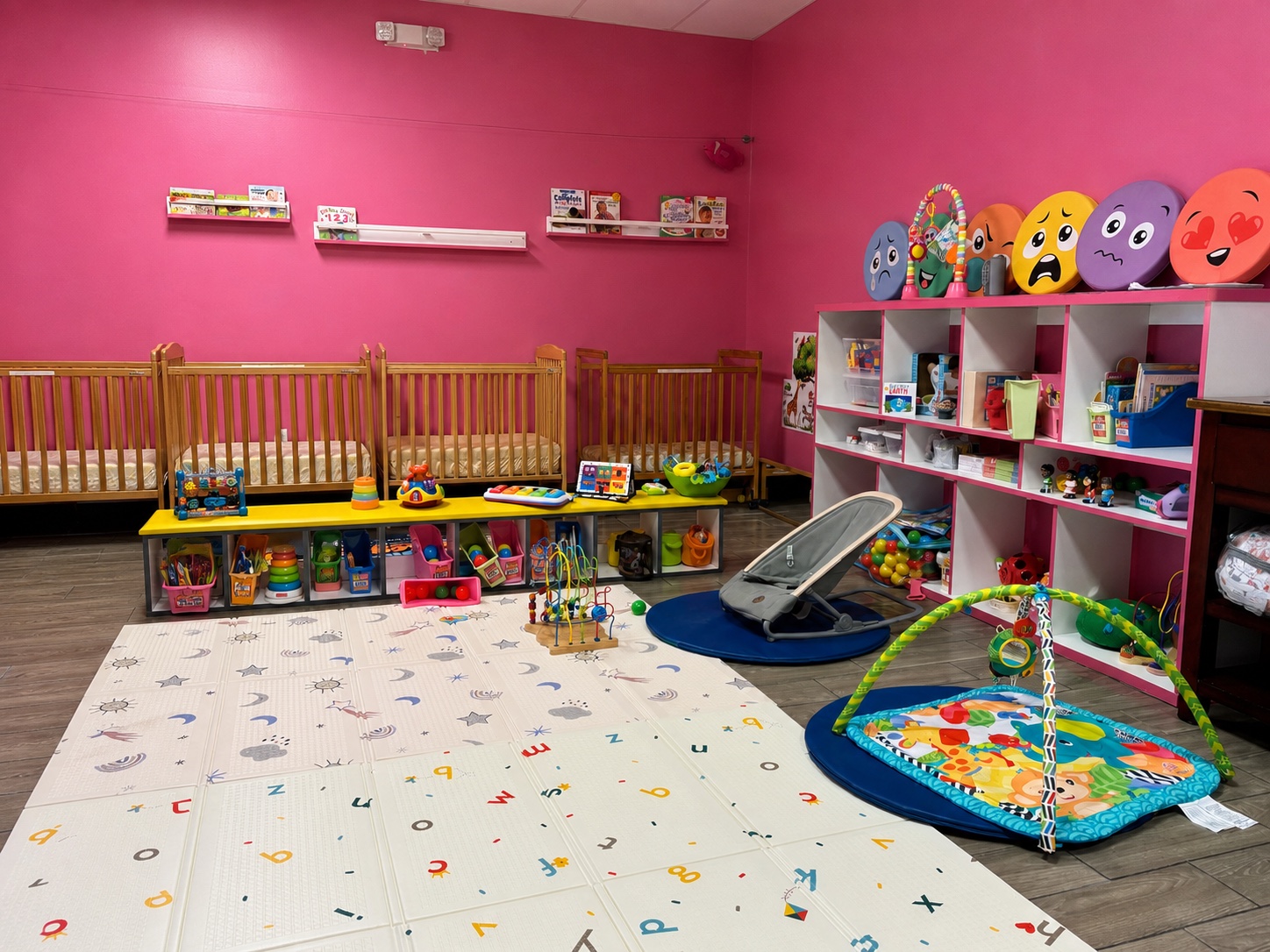 Bright pink infant classroom with cribs, play mats, toys, and organized learning materials